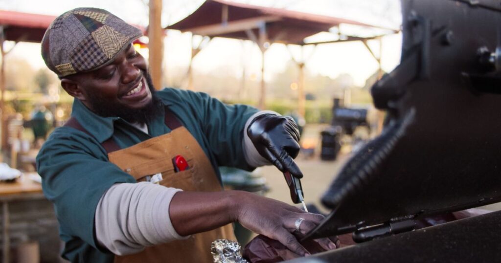 A man wearing an apron cooks on a grill, demonstrating his expertise in the BBQ competition series.