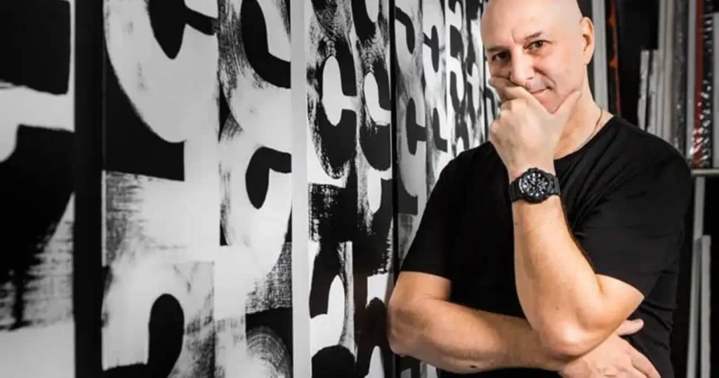 A bald man in a black shirt leans against a wall, representing Eric Haze's educational background and professional demeanor.