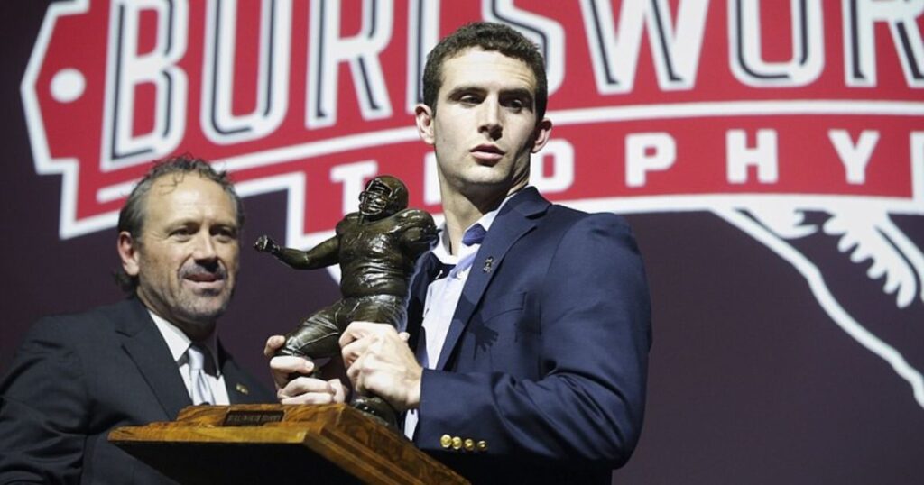 A man in a suit proudly holds a trophy in front of a sign, symbolizing Marty Burlsworth's success and achievements.